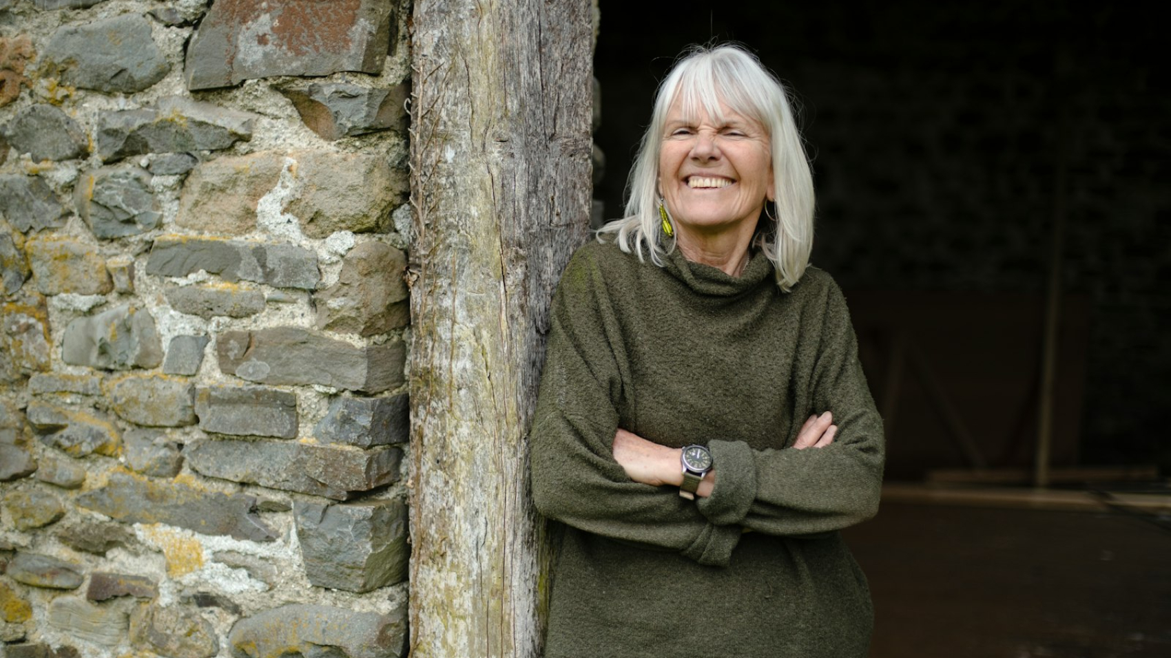 A smiling woman with grey hair leaning on a stone wall, confident and well through menopause